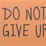 Cardboard sign reading 'Do Not Give Up' placed on green turf, symbolizing perseverance.